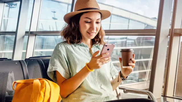 Kobieta w kapeluszu siedzi w poczekalni lotniskowej, trzymając telefon i kawę.
